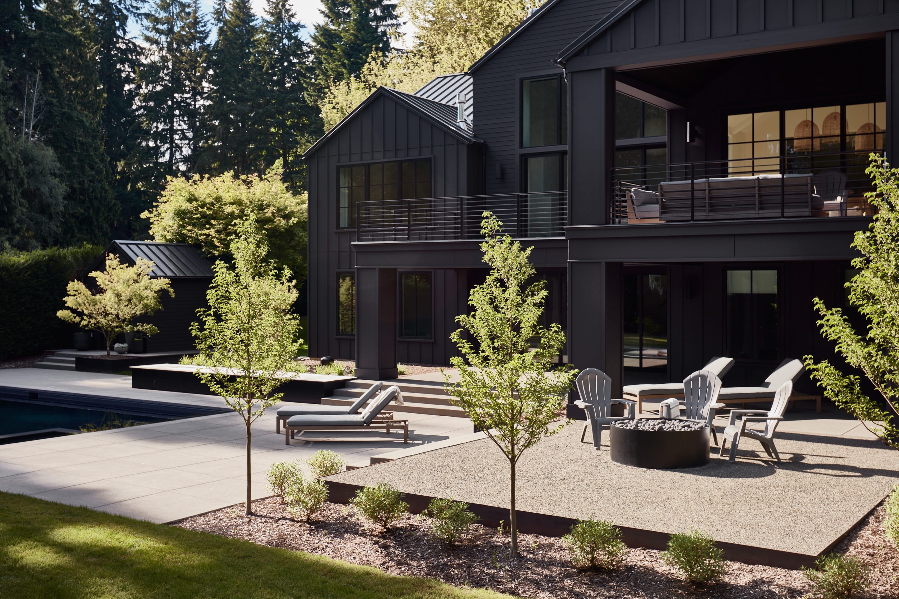 Modern black house with a patio featuring lounge chairs, a fire pit surrounded by Adirondack chairs, young trees, and a swimming pool.