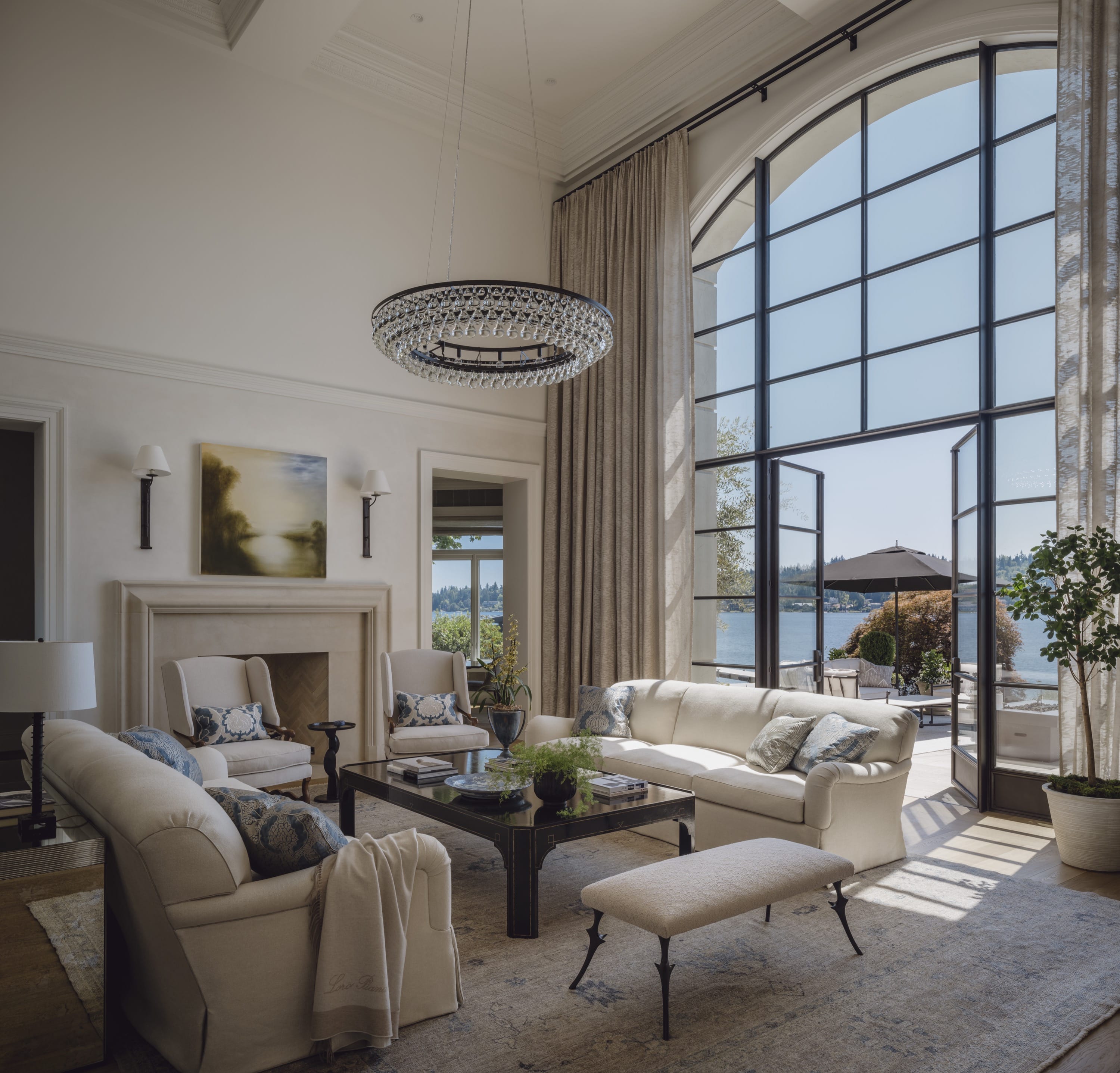 Elegant living room with beige sofas and armchairs, a black coffee table, large arched windows, and a view of a patio and lake.