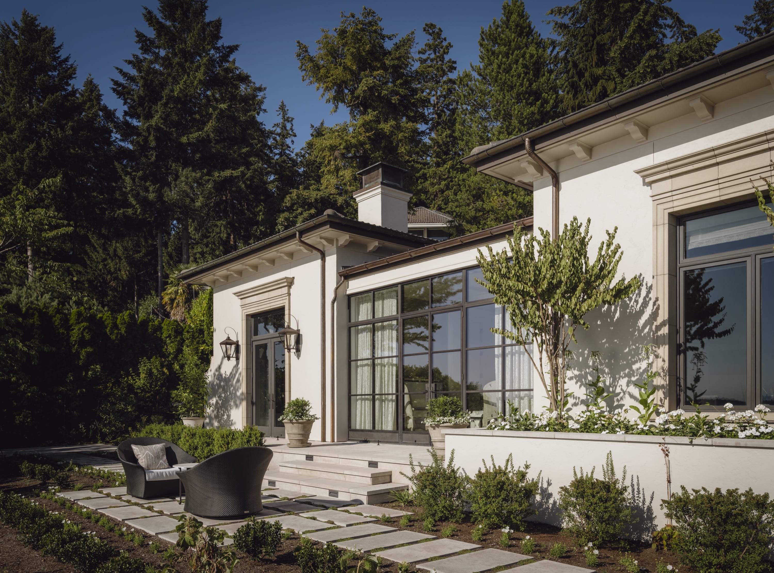 Modern house exterior with large windows, potted plants, and outdoor seating on a stone patio surrounded by greenery.