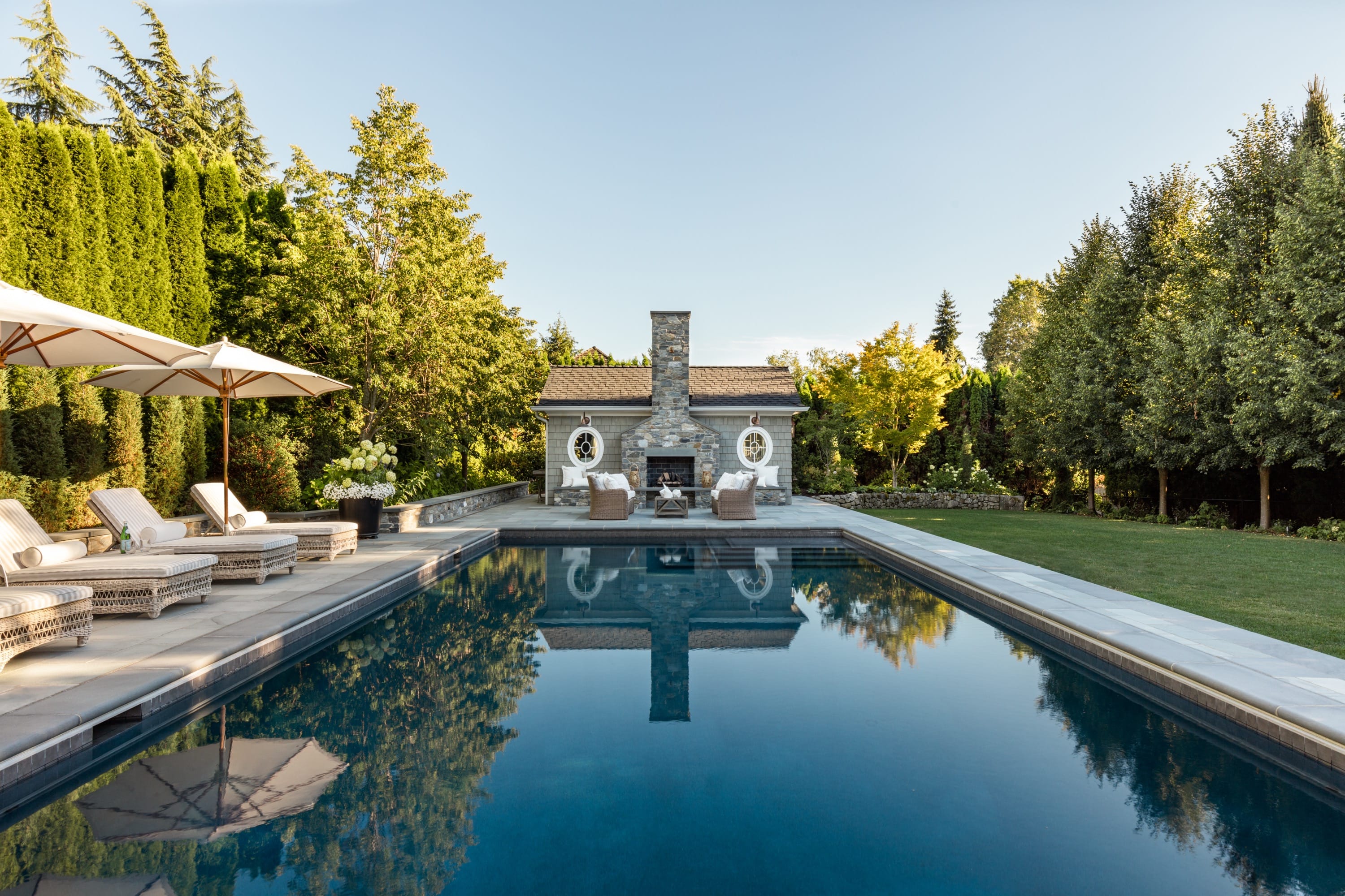 Elegant outdoor pool with lounge chairs and umbrellas on the left, reflecting trees and a small stone building with a chimney at the far end.