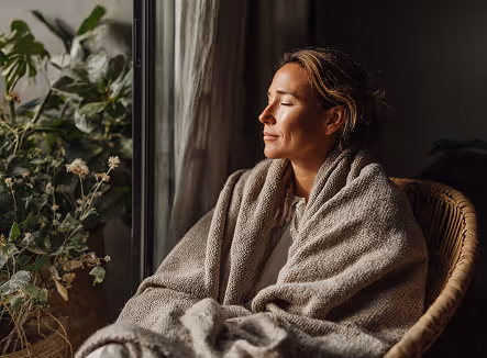 Woman wrapped in a cozy gray blanket sitting in a wicker chair by a window with plants nearby, eyes closed and enjoying natural light.