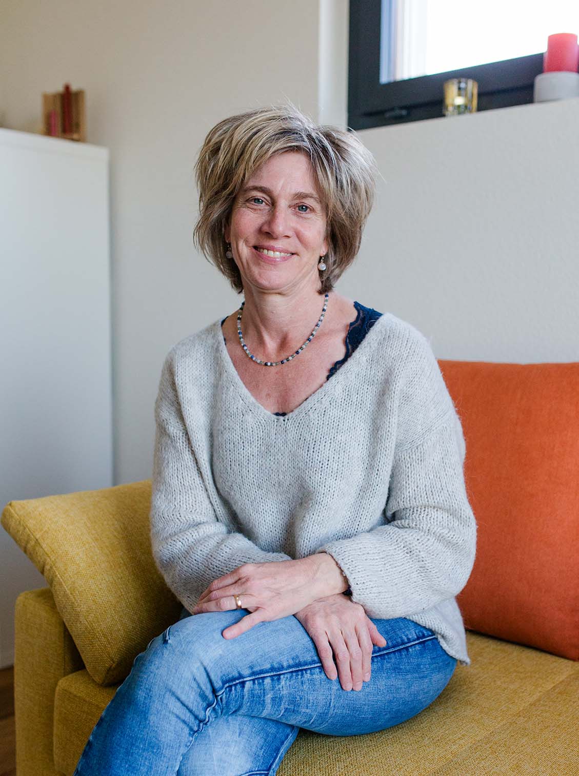 Smiling woman with short blonde hair wearing a gray sweater and blue jeans sitting on a yellow couch with an orange pillow.