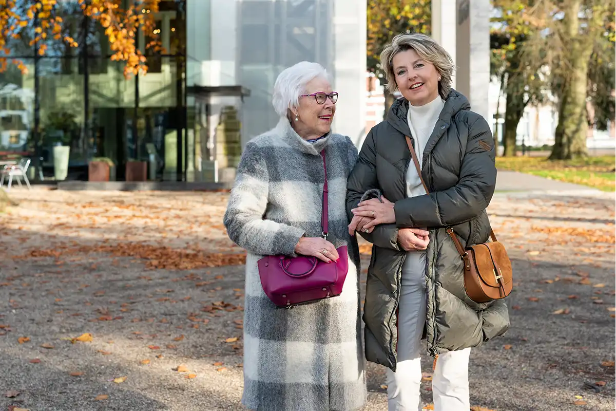 Oudere vrouw wandelt arm in arm met dochter in herfstomgeving. Complete uitleg over de Wet langdurige zorg en hoe u deze aanvraagt.