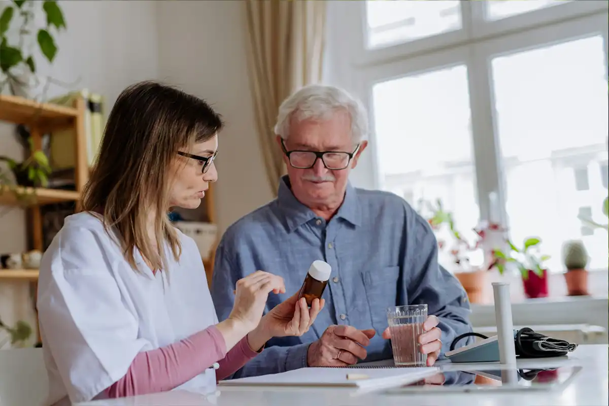 Vrouw helpt oudere man met medicatie aan keukentafel. Complete gids over dementie: herkenning, omgang en beschikbare zorg.