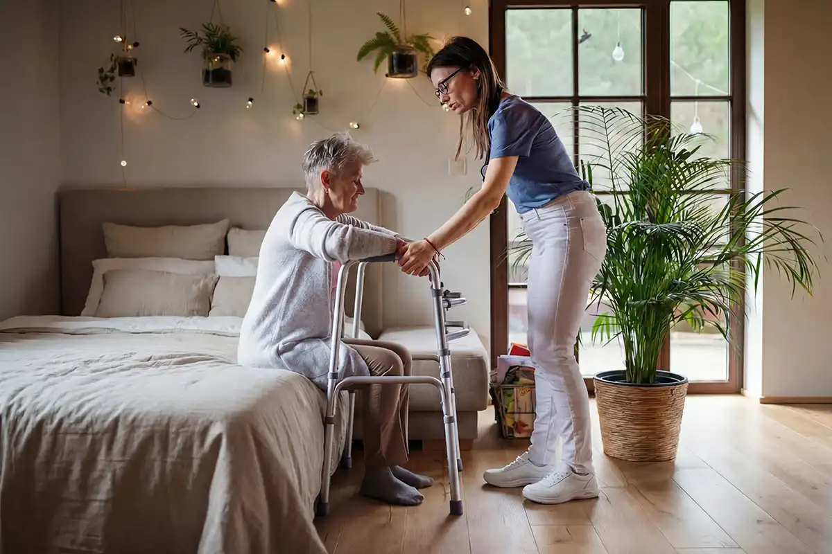 Zorgverlener helpt oudere vrouw met looprek uit bed in huiselijke slaapkamer. Uitleg over het persoonsgebonden budget en hoe u dit aanvraagt.