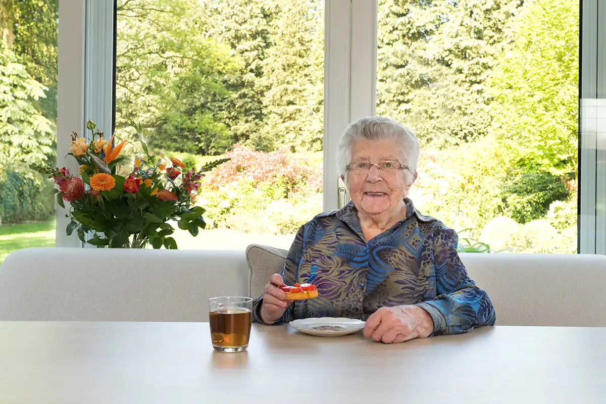 Oudere dame geniet van thee en koekje aan tafel met uitzicht op groene tuin. Uitleg over wat thuiszorg inhoudt en welke vormen er zijn.