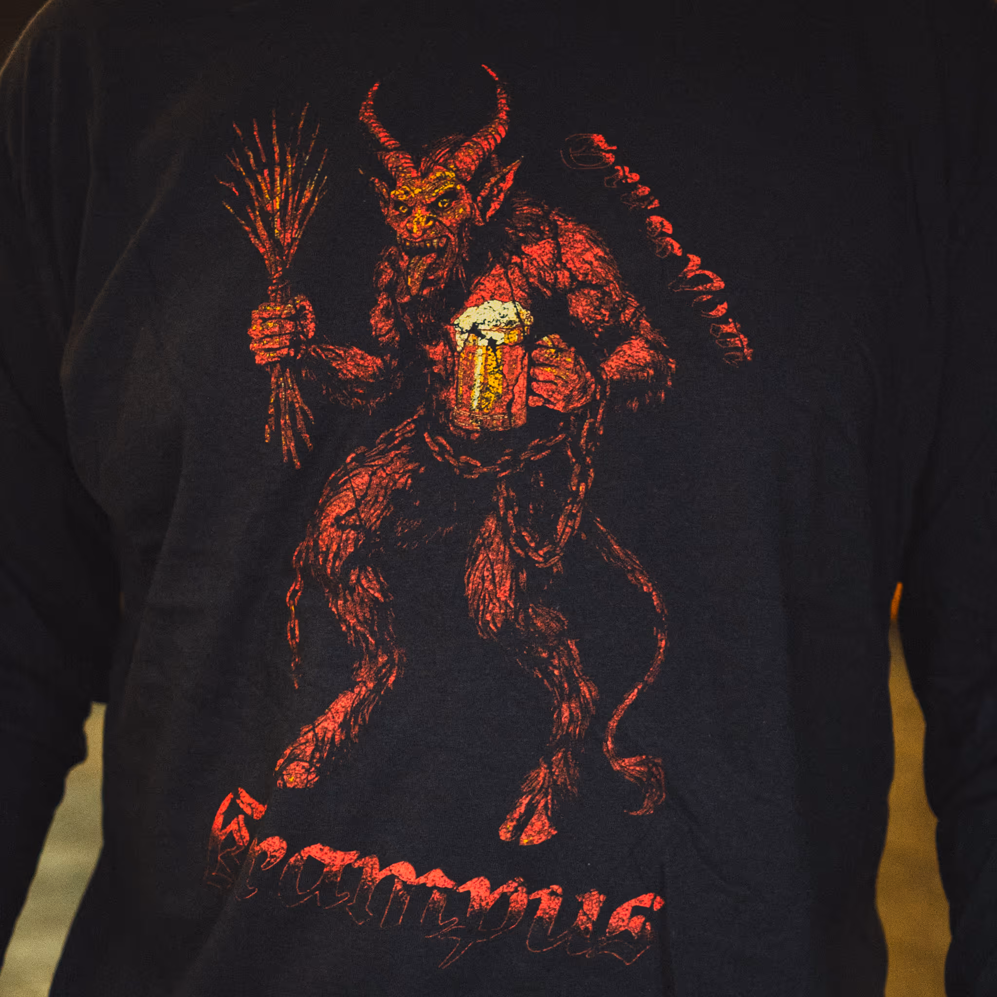 Black shirt featuring a red Krampus figure holding a bundle of sticks and a frothy beer mug, with stylized text below.