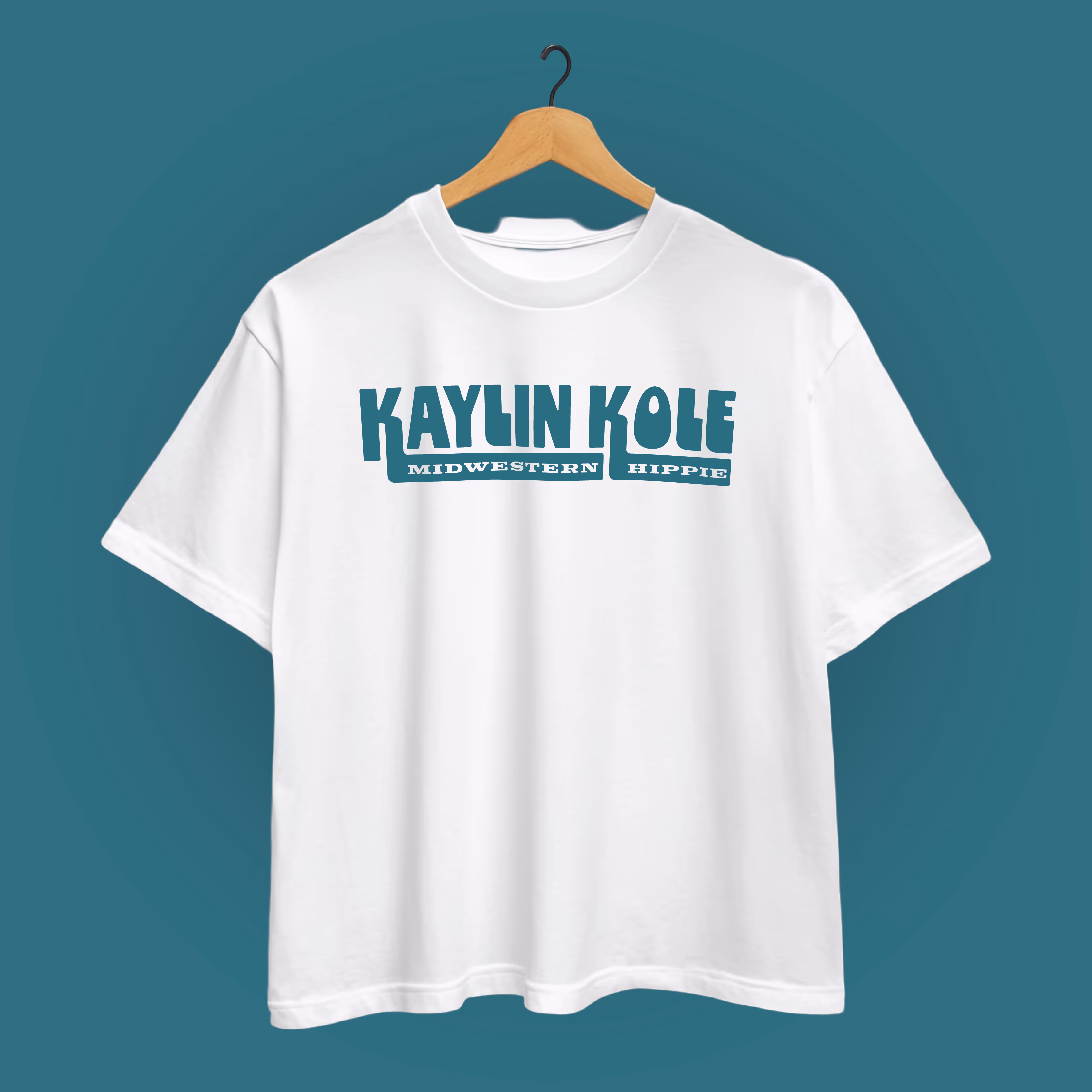 White t-shirt on wooden hanger with teal text saying 'KAYLIN KOLE' and 'MIDWESTERN HIPPIE' below it.