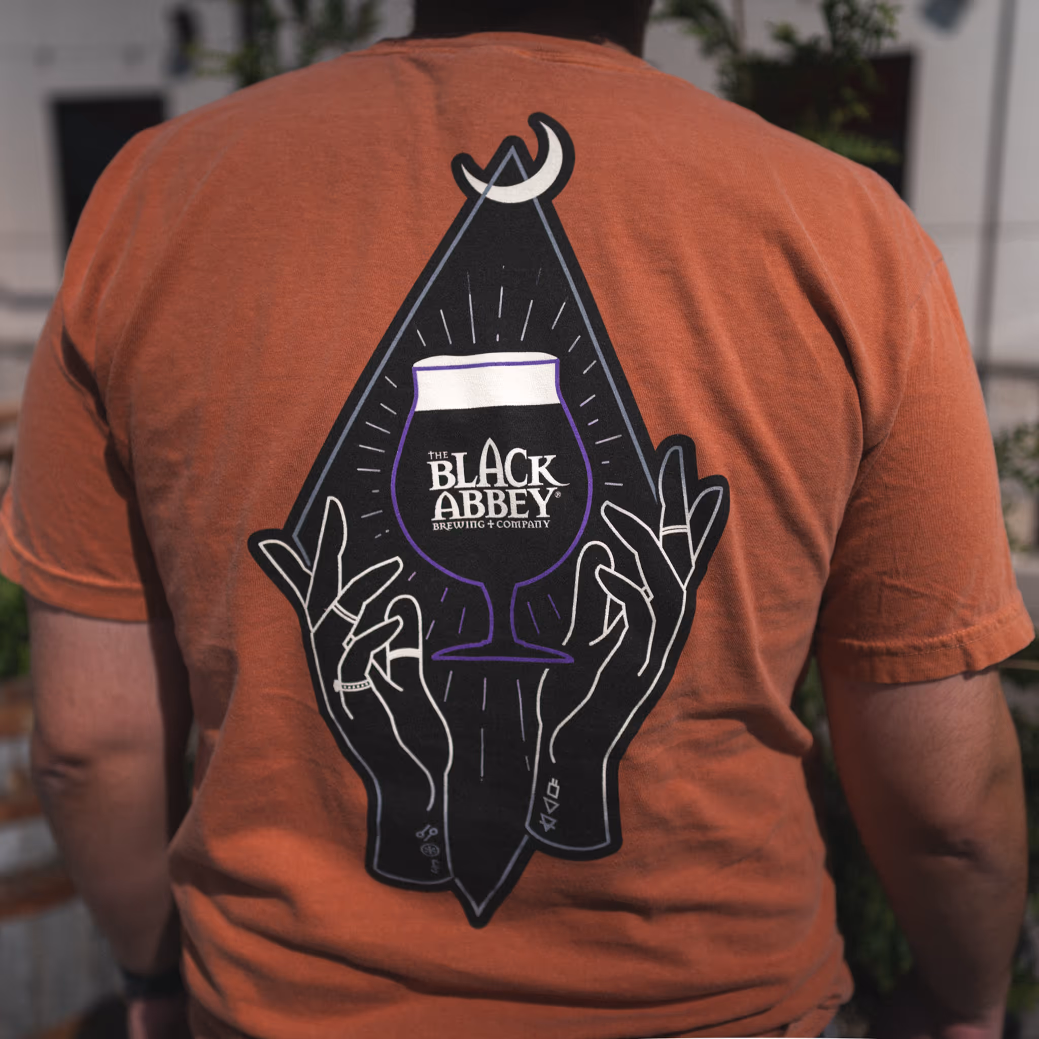 Back of a person wearing an orange shirt with a graphic of two hands holding a beer glass labeled The Black Abbey Brewing + Company under a crescent moon.