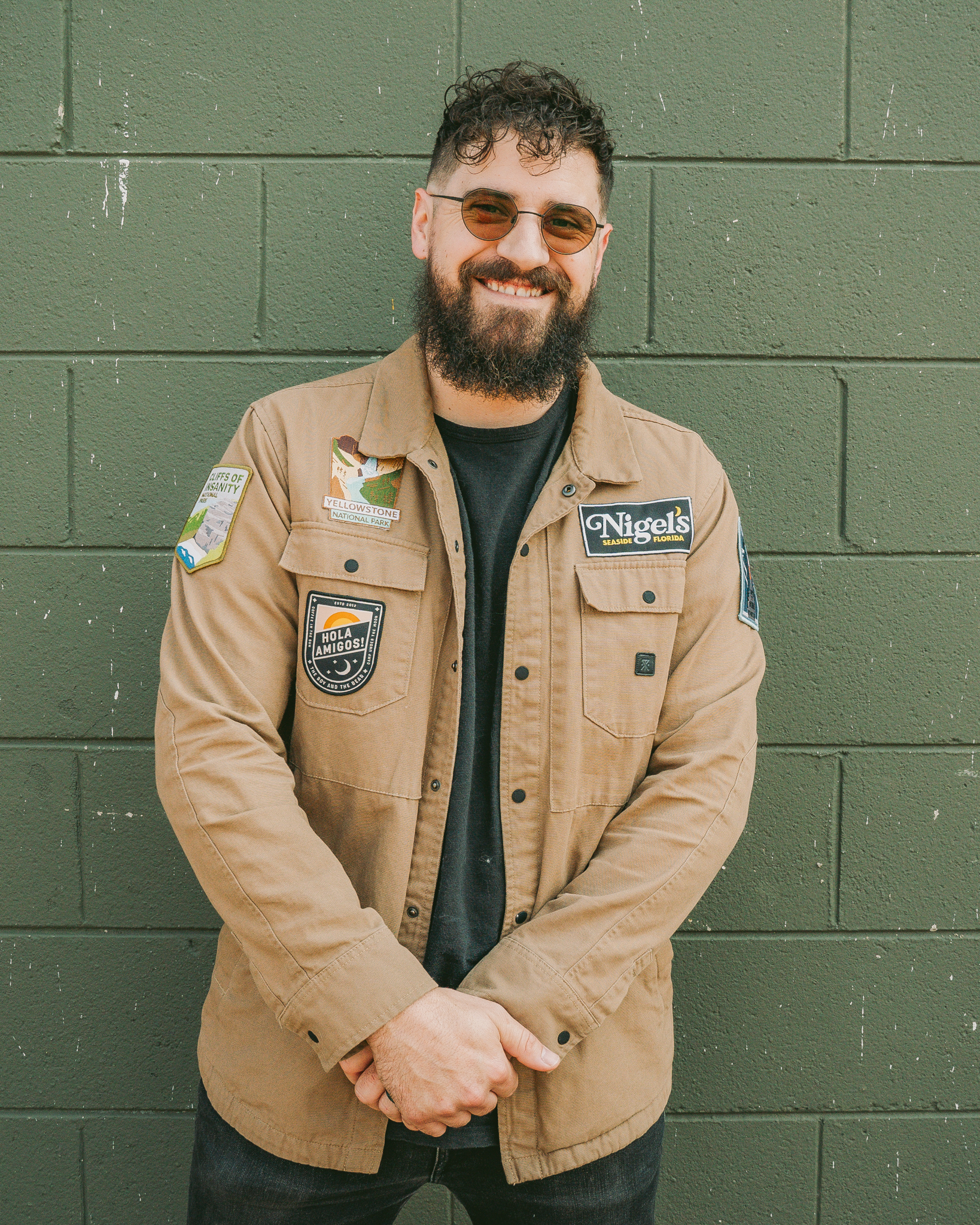 Smiling man with curly hair and beard wearing round sunglasses and a tan jacket with travel patches standing in front of a green cinder block wall.