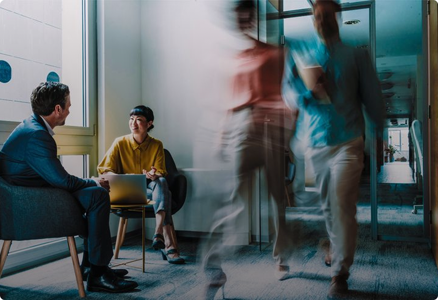 employees walking through an office space whilst others sit and have a casual meeting over a laptop