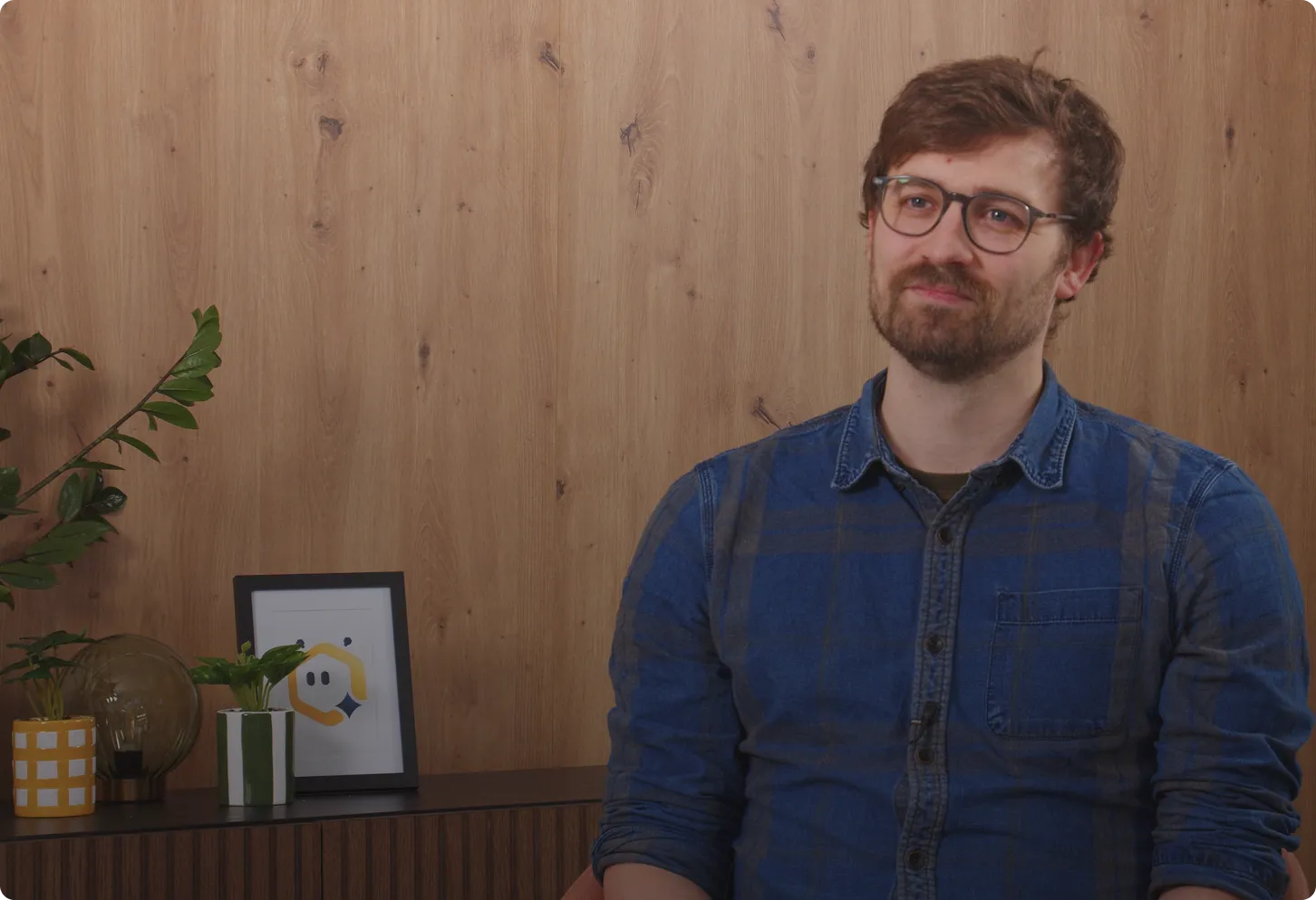 Man with glasses and beard wearing a blue shirt sitting in front of a wood-paneled wall with small plants and a framed picture on a sideboard.