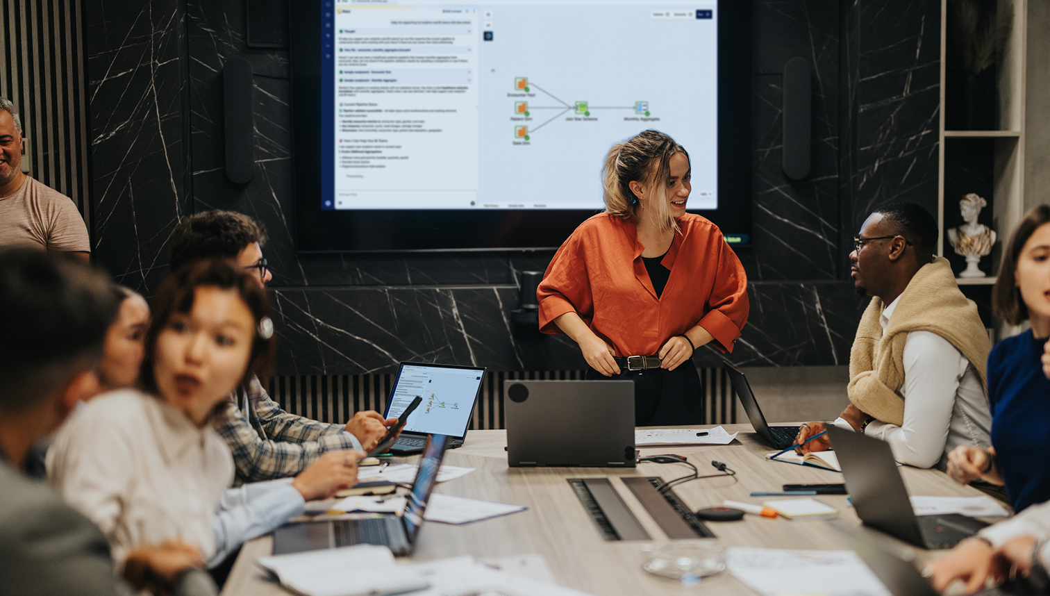 People gathered around a conference table with laptops, discussing in front of a screen showing a workflow.
