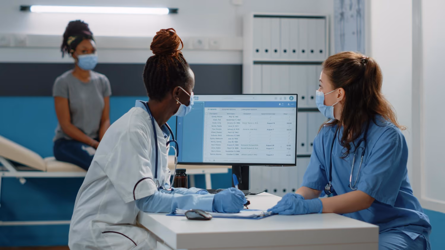 Two clinical trial professionals in medical scrubs performing Site Management of site patient information displayed on a computer screen in a clinic room with a seated patient in the background.