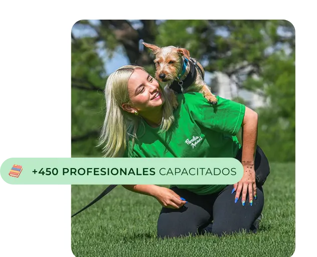 Mujer con camiseta verde arrodillada en césped mientras carga un perro pequeño sobre sus hombros, ambos al aire libre.