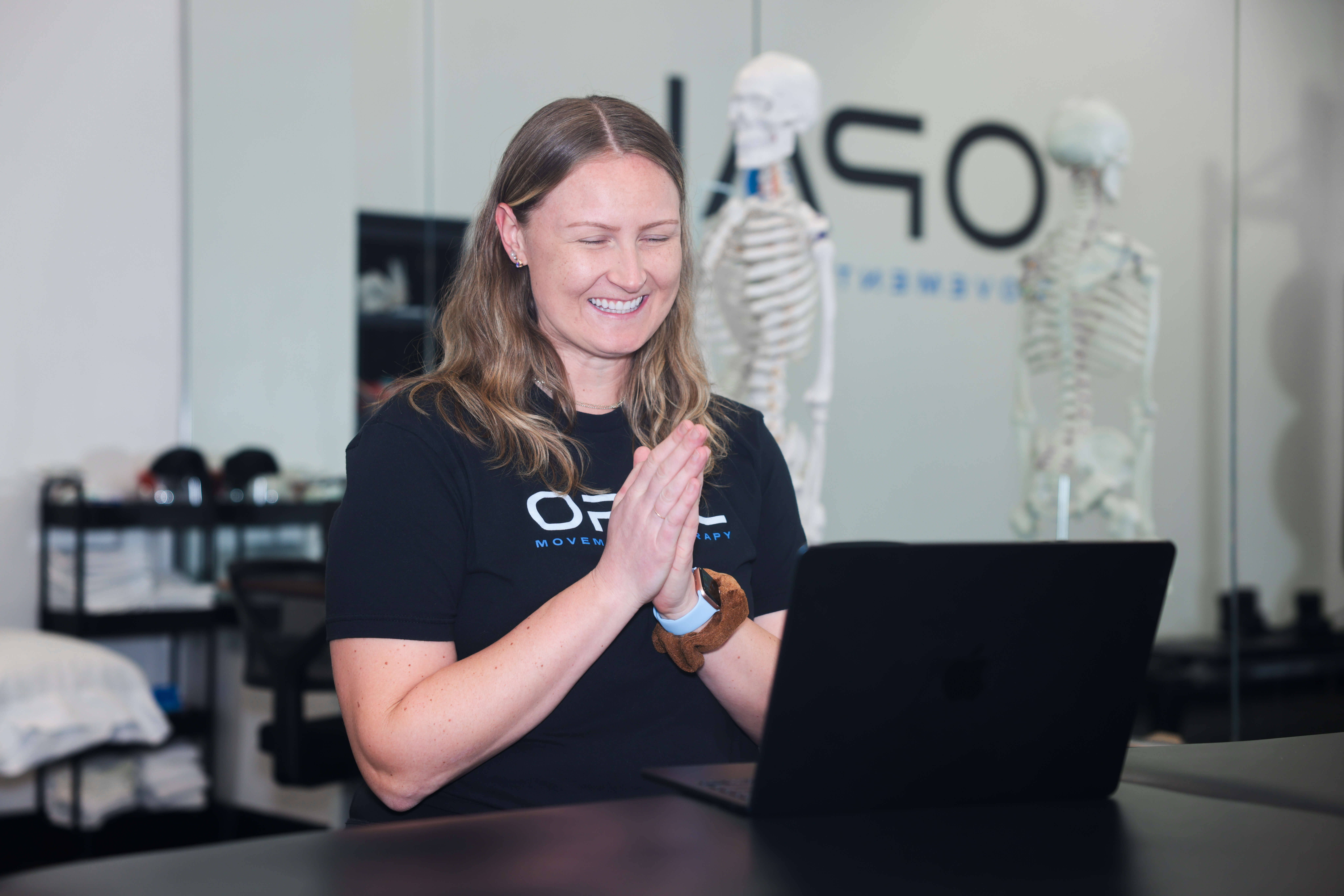Smiling woman with folded hands looking at a laptop in a therapy or fitness studio with skeleton models in the background.