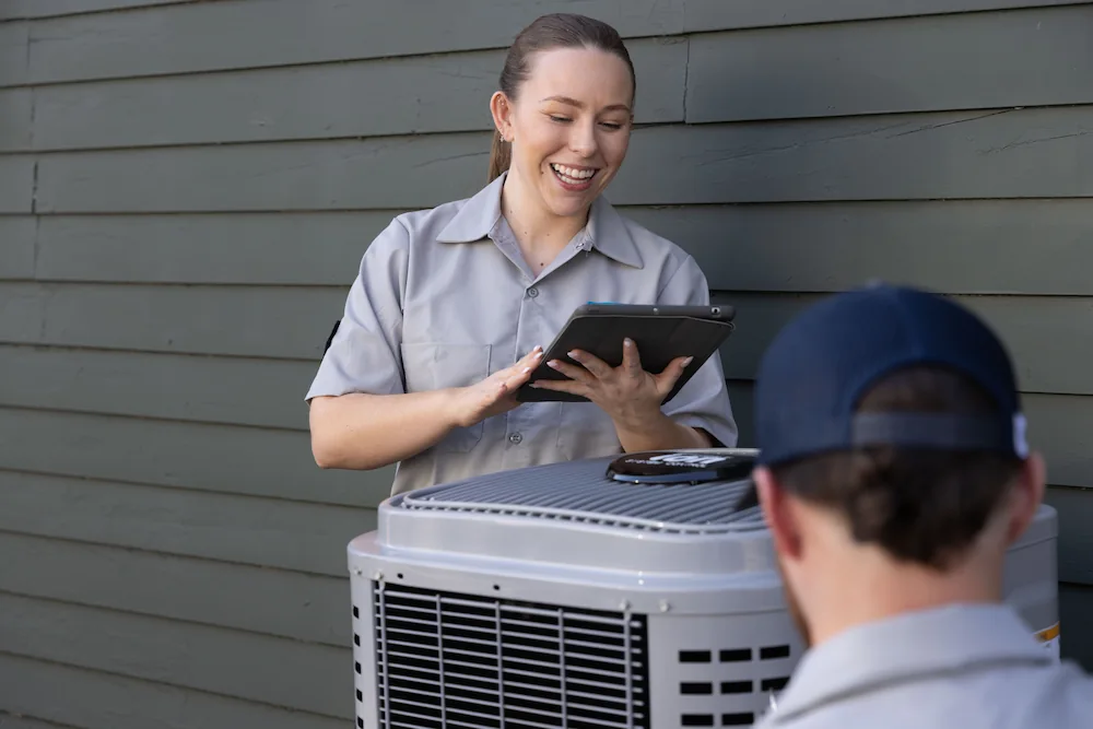 Two HVAC technicians working on an air conditioning unit outside a building, with one technician using a tablet and smiling.