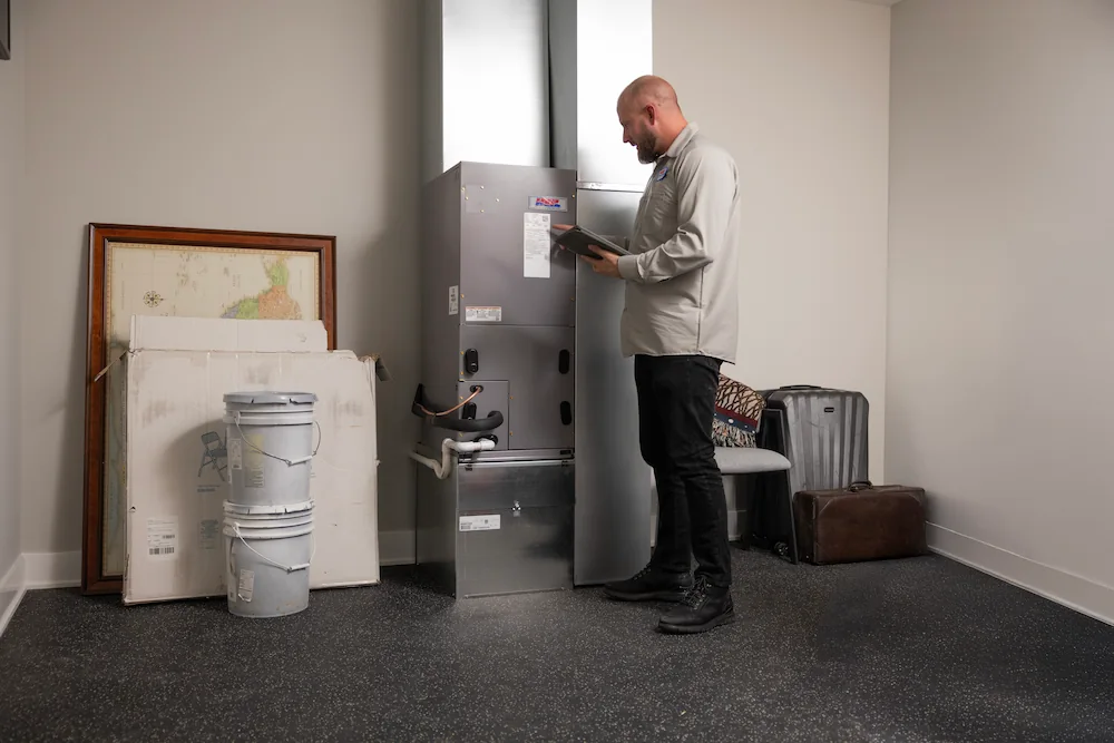 Technician inspecting an HVAC unit inside a room with tools and storage items nearby.