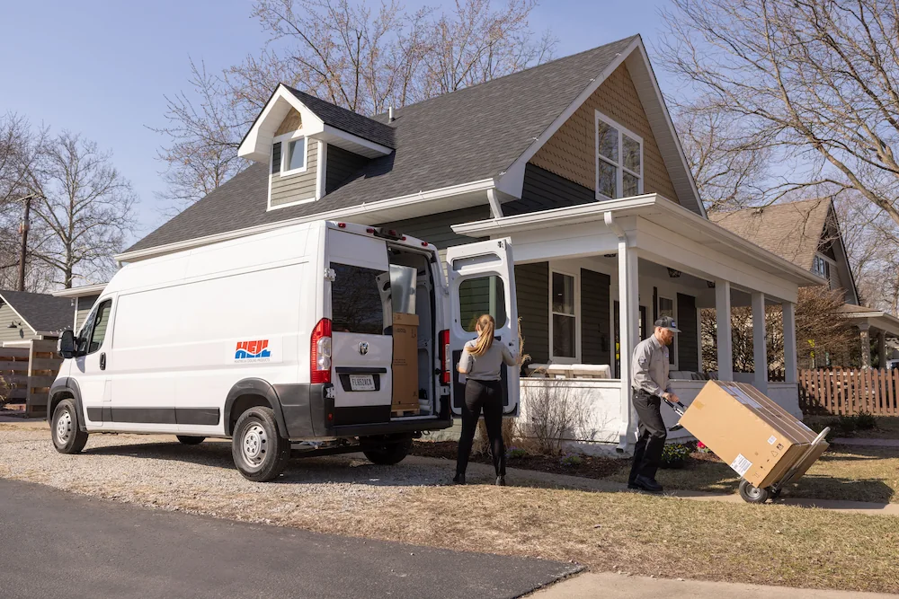 Two people unloading large boxes from a white delivery van in front of a house with a porch.