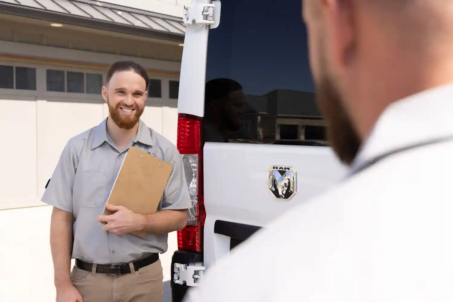 Smiling delivery man holding a clipboard standing next to a white Ram van outdoors.