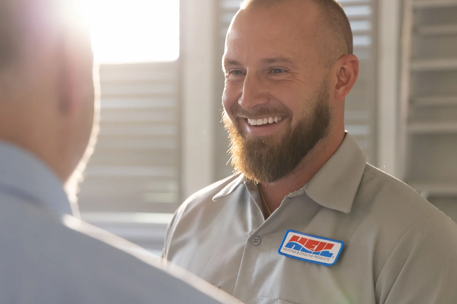 Smiling man with a beard wearing a HEIL Heating & Cooling Products uniform shirt engaged in conversation.
