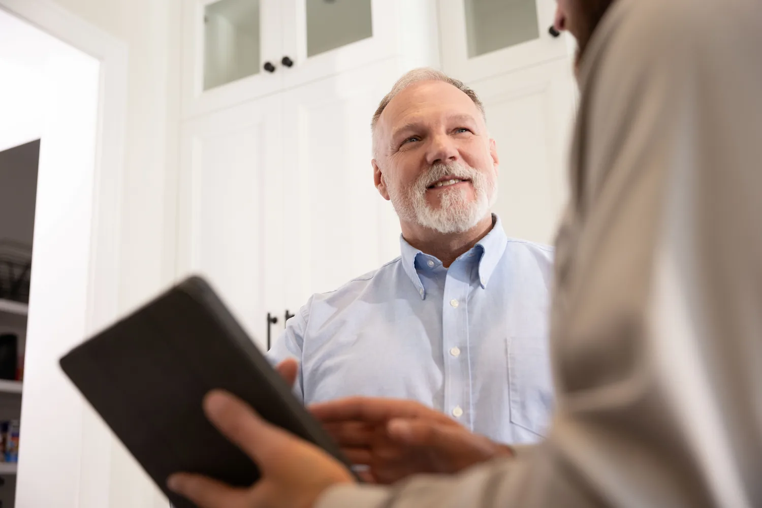 Smiling older man with white beard wearing a light blue shirt talking to another person holding a tablet.