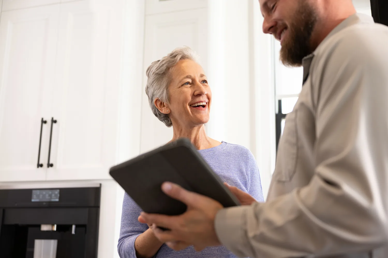 Smiling older woman talking to a man holding a tablet in a bright kitchen.