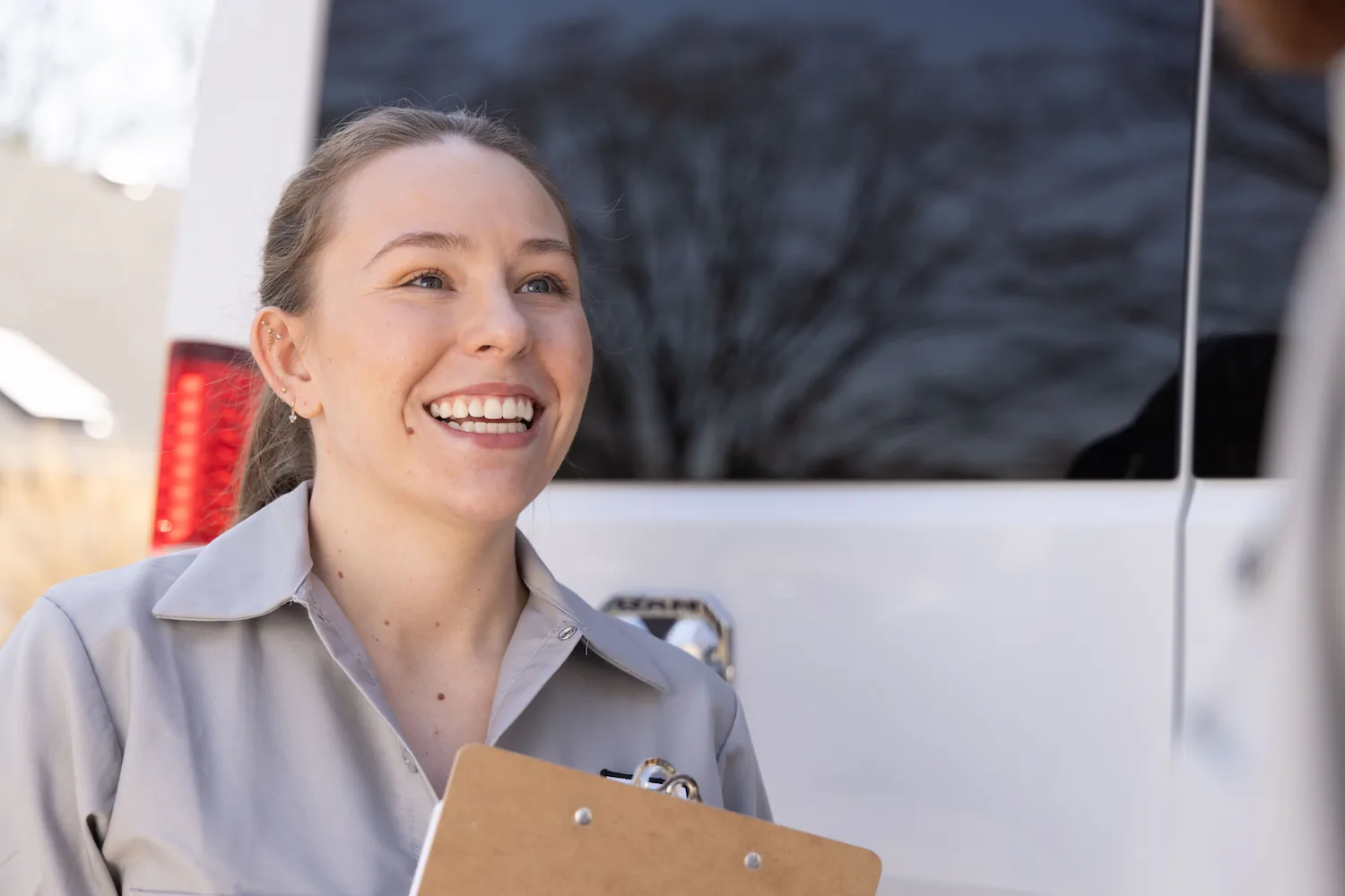 Smiling woman in a light gray uniform holding a clipboard, standing near a white vehicle.