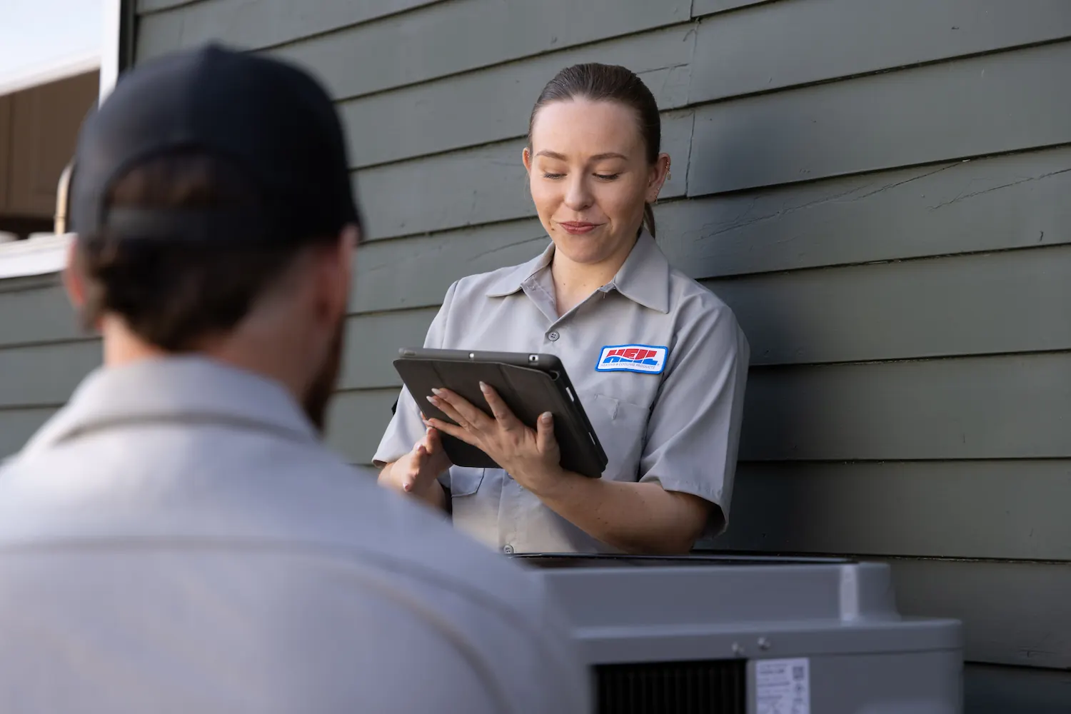 Technician in a gray uniform using a tablet next to an outdoor HVAC unit with another technician in the foreground.