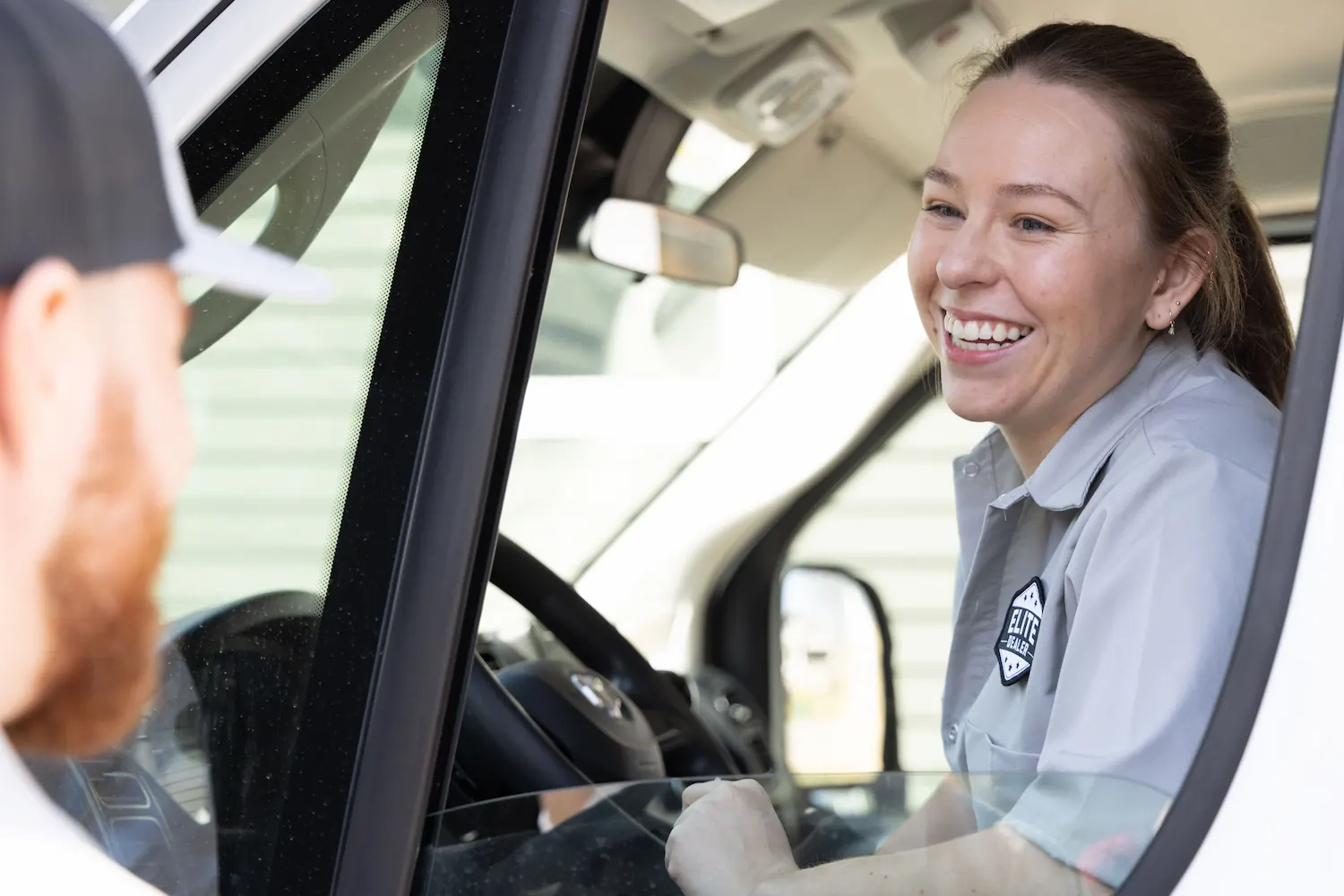 Smiling woman in a gray uniform shirt talking to a man with a beard sitting in the driver's seat of a vehicle.