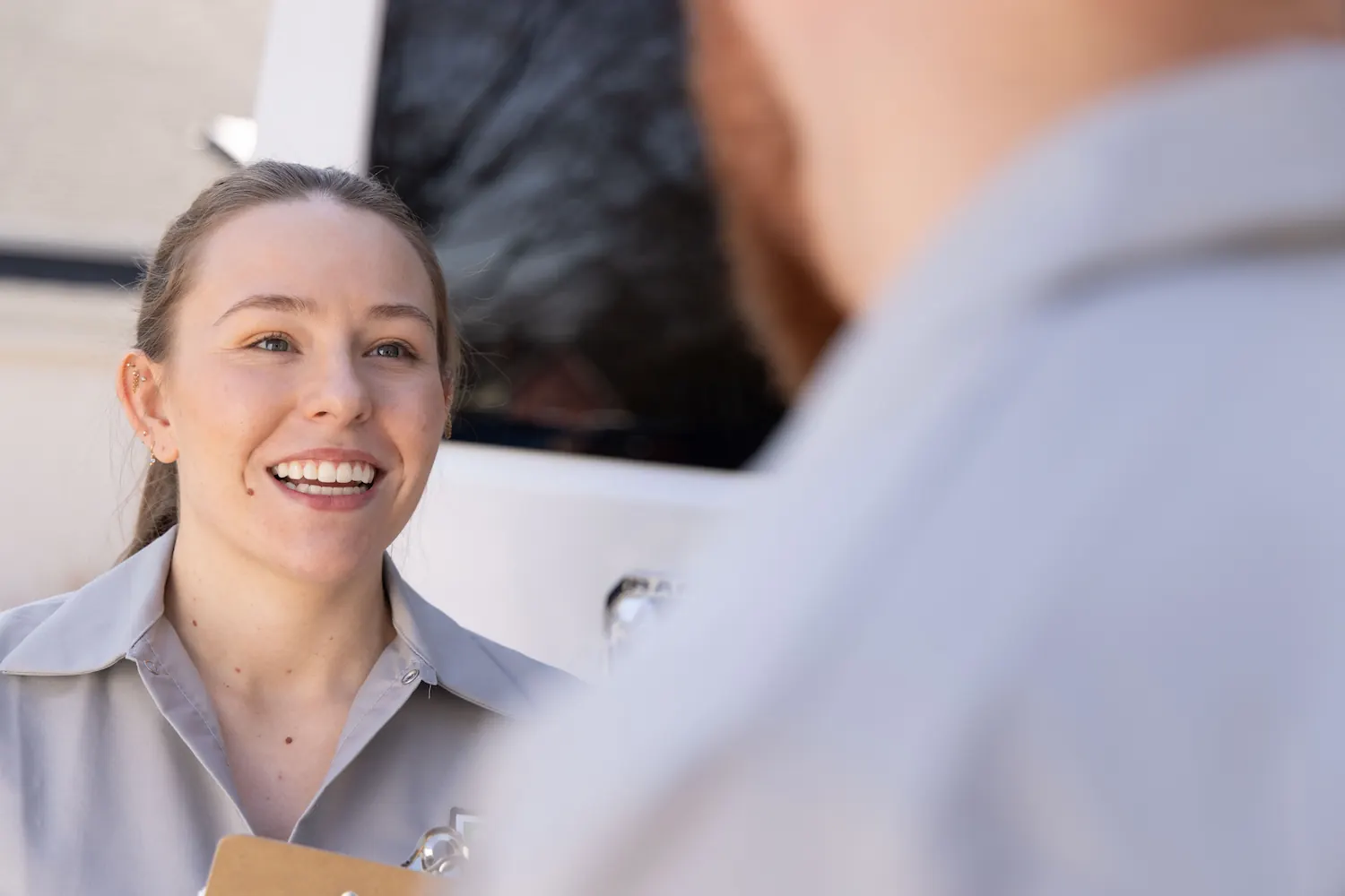Smiling woman in a gray uniform holding a clipboard talking to a person outdoors.