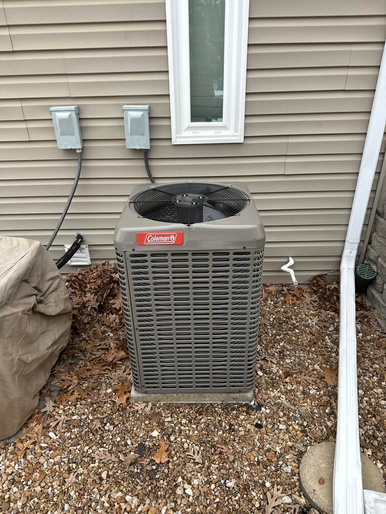 Outdoor Coleman air conditioning unit installed on a gravel bed beside a house with beige siding and a small window.