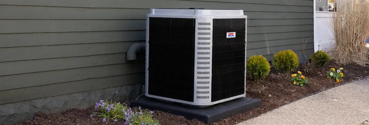 Outdoor air conditioning unit installed next to a gray house with white-trimmed windows and landscaped garden bed.