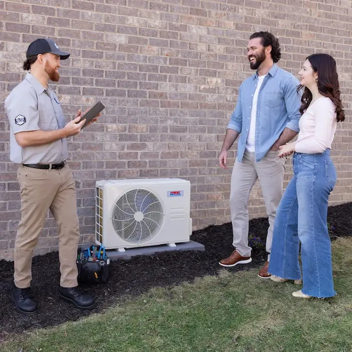 HVAC technician explaining ductless heat pump installation to a smiling couple outside their brick house.