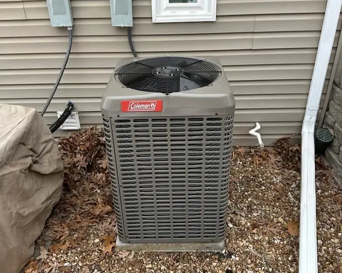 Outdoor Coleman air conditioning unit installed on a gravel bed beside a house with beige siding and a small window.