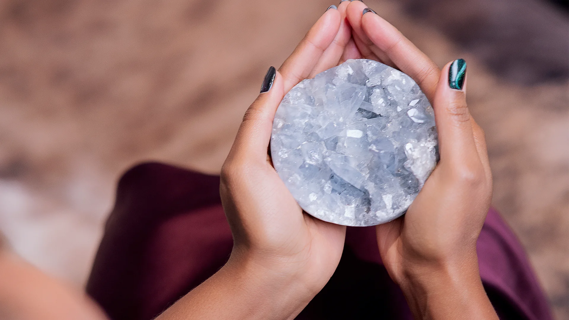 Hands with black and green nail polish gently holding a round cluster of clear quartz crystals.