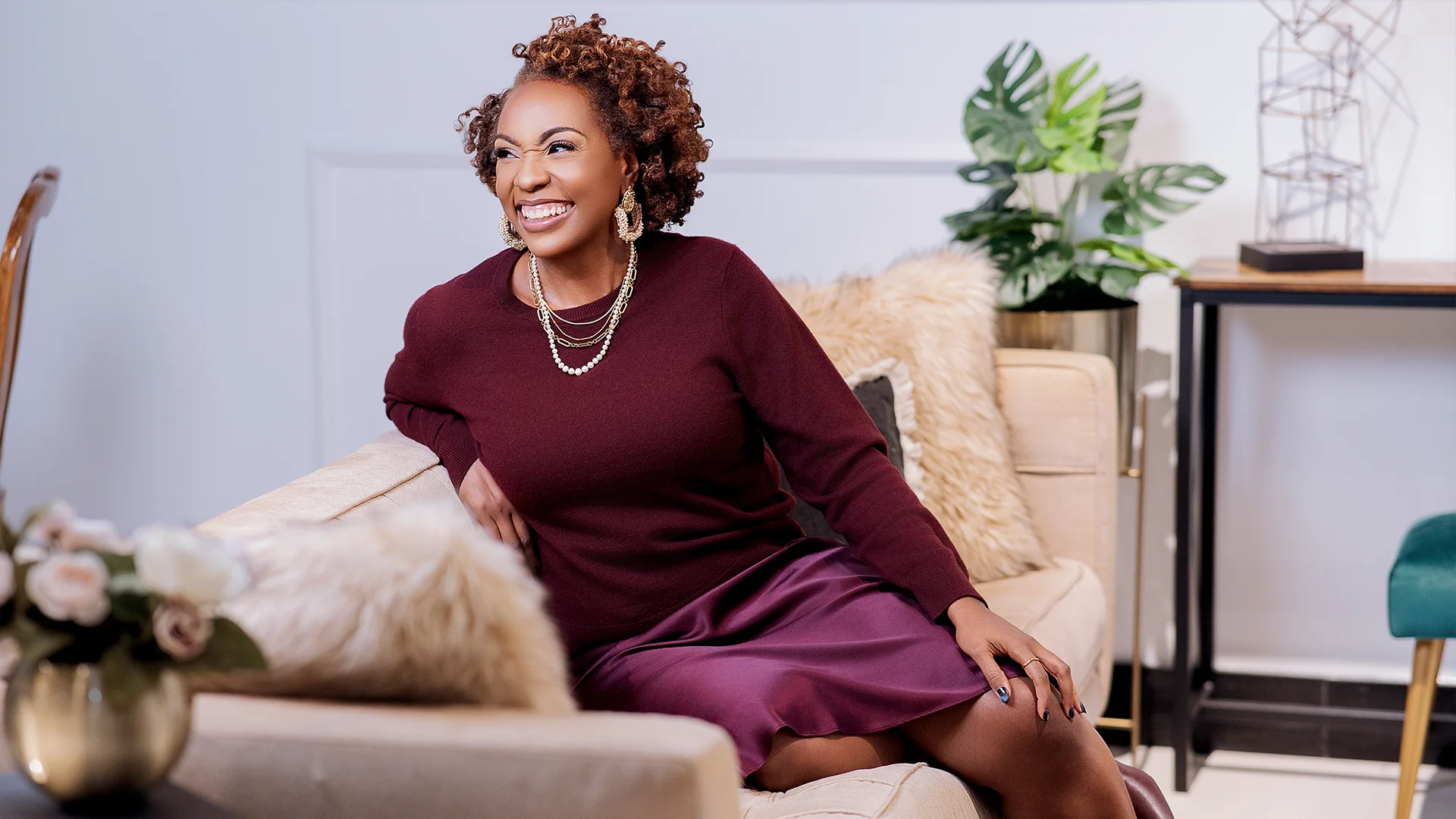 Smiling woman with curly hair sitting on a beige couch wearing a burgundy sweater and a purple skirt.