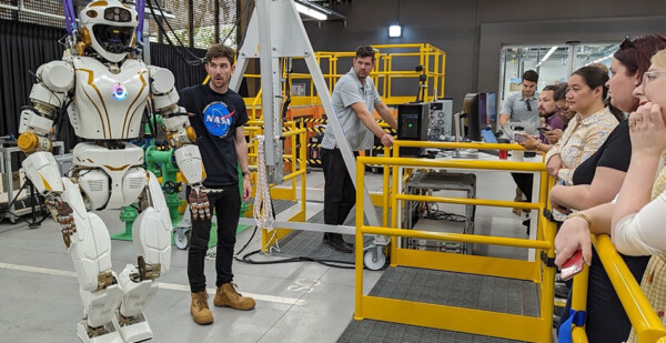 Man in NASA shirt stands with humanoid robot while conducting demonstration to company representatives (Woodside)