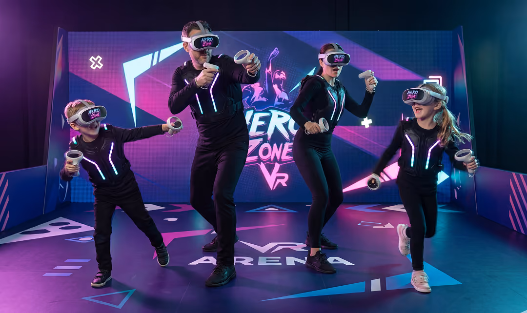 A family of four wearing VR headsets and glowing suits playing an interactive game in a VR arena with a colorful Hero Zone VR backdrop.