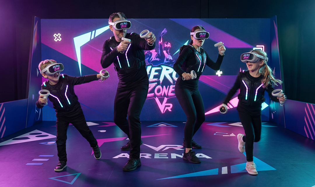 A family of four wearing VR headsets and glowing suits playing an interactive game in a VR arena with a colorful Hero Zone VR backdrop.