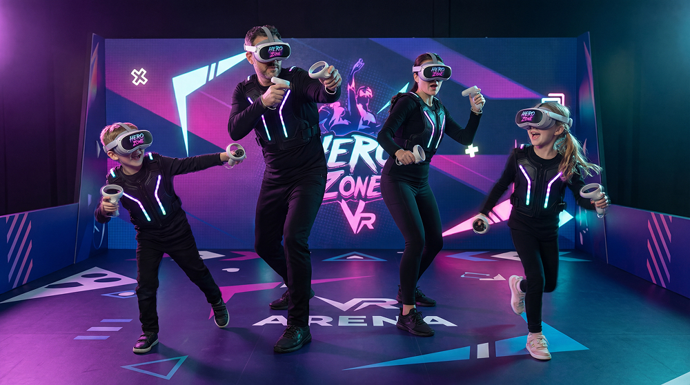 Four people in black suits with glowing stripes wearing HERO ZONE VR headsets and holding controllers in a VR gaming arena.