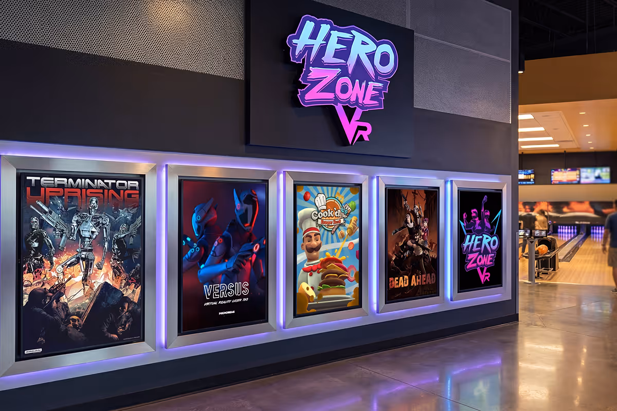 Wall display of five framed VR game posters under a glowing Hero Zone VR sign in an entertainment venue with a bowling alley in the background.