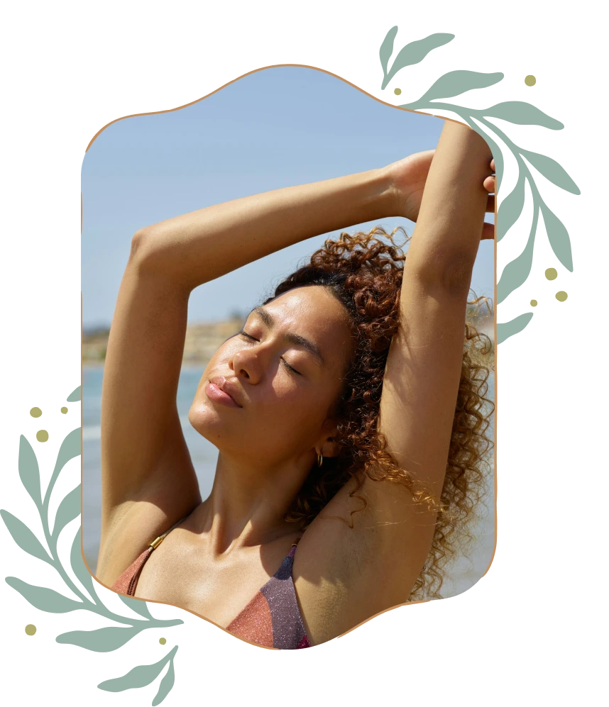 Woman with curly hair in a swimsuit stretching arms above head with eyes closed on a sunny beach.