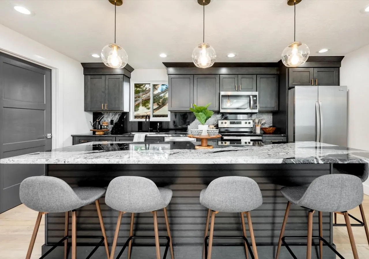 Modern kitchen with dark gray cabinets, stainless steel appliances, granite countertop island, and four gray upholstered bar stools.