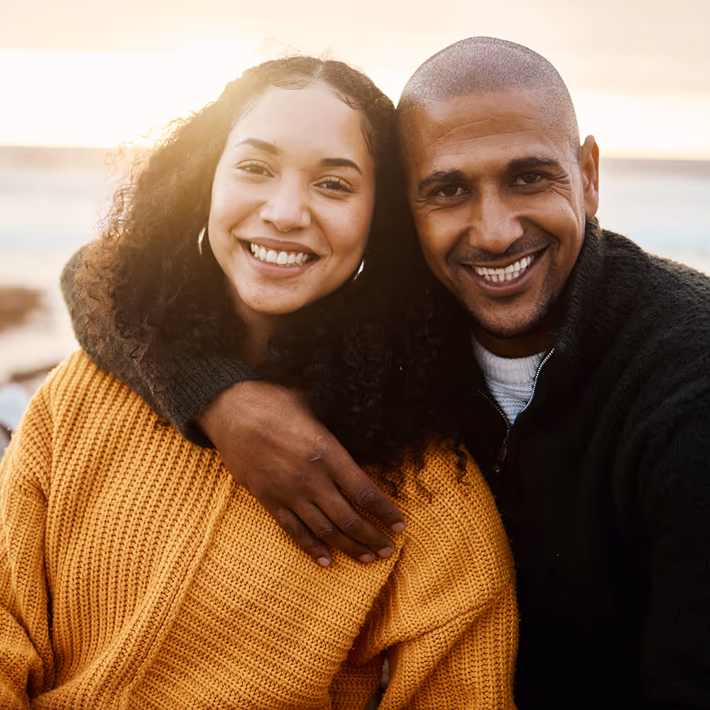 Smiling couple outdoors with man's arm around woman's shoulder during sunset.