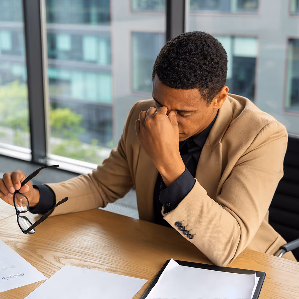 Stressed man in a beige jacket sitting at a desk holding glasses and rubbing his eyes.