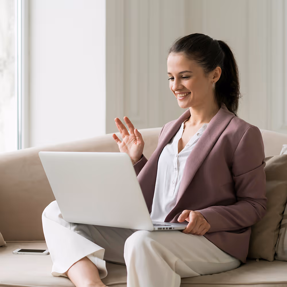 Smiling woman sitting on a couch using a laptop and waving during a video call.