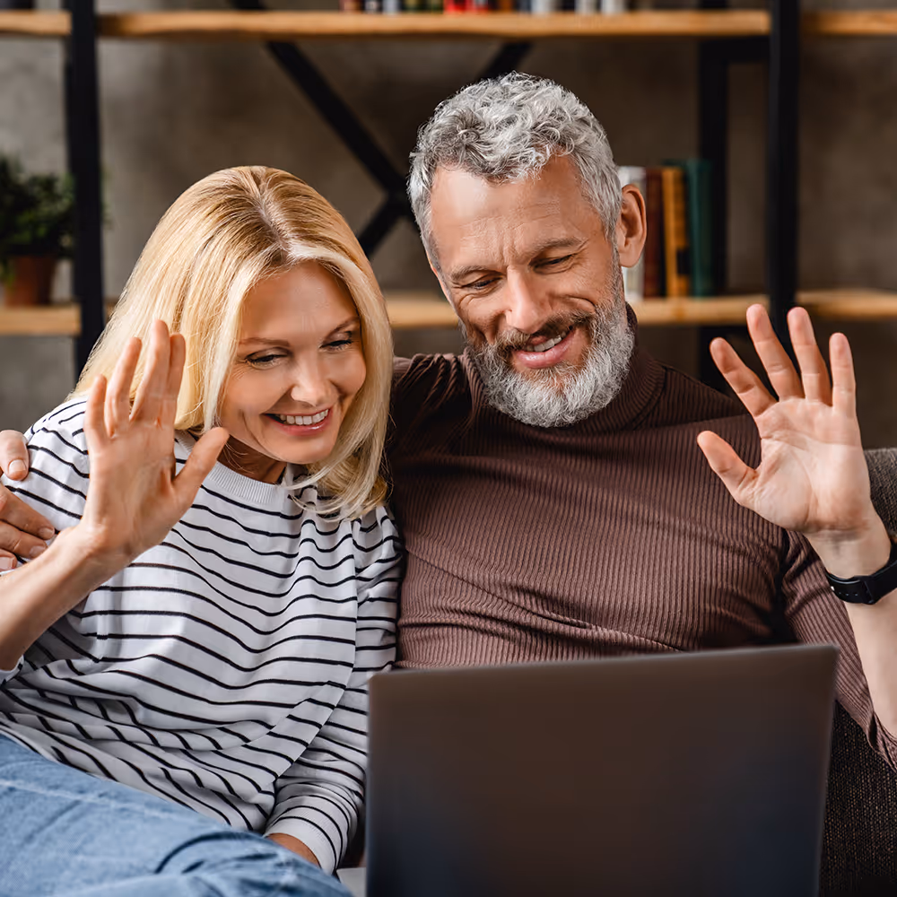 Smiling middle-aged couple sitting on a couch waving at a laptop screen during a video call.