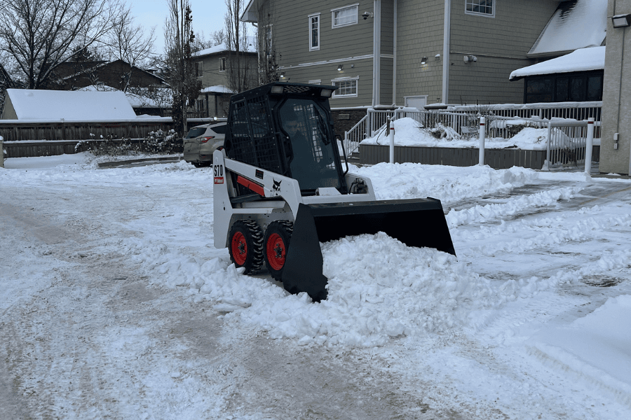 An image of a white and orange skid steer plowing snow as part of Badger Landscaping's commercial snow removal Edmonton Services. They are clearing the snow out of a parking lot with the skid steer.