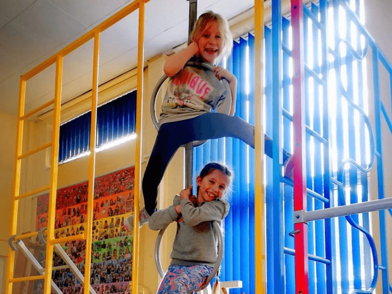 Two girls smiling on a climbing frame in a sports hall.